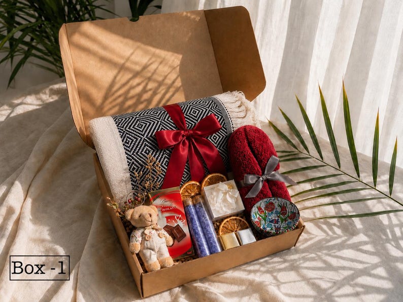 K&ouml;nnte beinhalten: Eine Geschenkschachtel aus Karton mit einer schwarz-wei&szlig; gemusterten Decke mit roter Schleife, einem kleinen Teddyb&auml;ren, einer roten Schokoladentafel, zwei violetten Badesalzr&ouml;hrchen, getrockneten Orangenscheiben und einem roten Handtuch.