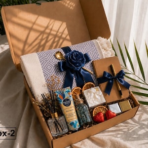 K&ouml;nnte beinhalten: Eine Geschenkbox mit der Aufschrift Box-2, die eine grau-wei&szlig; gemusterte Decke mit dunkelblauem Band und Rose, ein braunes Notizbuch mit goldenem Stift, eine Handcreme, einen Glas-Diffusor, getrocknete Orangenscheiben und kleine Herzbonbons enth&auml;lt.