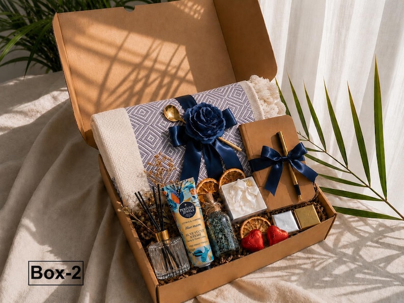 K&ouml;nnte beinhalten: Eine Geschenkbox mit der Aufschrift Box-2, die eine grau-wei&szlig; gemusterte Decke mit dunkelblauem Band und Rose, ein braunes Notizbuch mit goldenem Stift, eine Handcreme, einen Glas-Diffusor, getrocknete Orangenscheiben und kleine Herzbonbons enth&auml;lt.