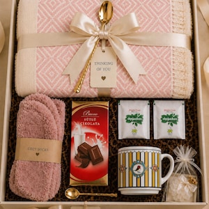 May include: A gift box containing a pink and cream diamond-patterned throw blanket, a pair of fuzzy pink socks, a bar of Buono milk chocolate, two Dogadan tea bags, a striped mug with a bird motif, two gold spoons, and a small floral sachet.