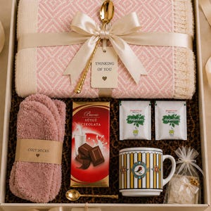 May include: A gift box containing a pink and cream diamond-patterned throw blanket, a pair of fuzzy pink socks, a bar of Buono milk chocolate, two Dogadan tea bags, a striped mug with a bird motif, two gold spoons, and a small floral sachet.