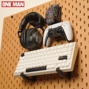 Puede incluir: Una exhibición de accesorios de juego, que incluye un teclado blanco, auriculares negros, un mando de juego blanco y negro y un ratón con un diseño colorido. Los artículos están montados en un panel perforado marrón.