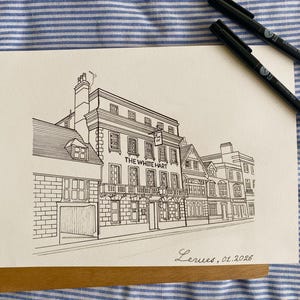以下が含まれることがあります： 「THE WHITE HART」の文字が入った建物の白黒インク画。窓や看板など、詳細な建築ファサードを描いた作品です。黒いペンが2本見えます。