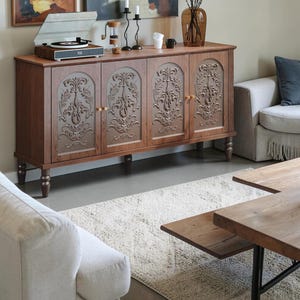 May include: A rectangular wooden sideboard featuring four cabinet doors with arched panels and intricate, raised floral carvings. The piece stands on six turned legs and includes small, round wooden knobs on each door for opening the storage compartments.