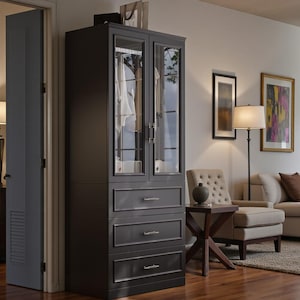 May include: A dark gray wardrobe with glass doors and three drawers. The wardrobe has a classic design with silver hardware and is filled with clothing. The wardrobe is in a well-lit room with a hardwood floor.