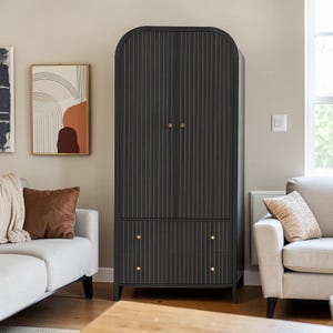 May include: A tall, black wooden wardrobe with a vertically striped design. The wardrobe has two doors with brass-colored knobs and two drawers with matching knobs. The rounded top and sleek design give it a modern look. The wardrobe is in a living room setting.