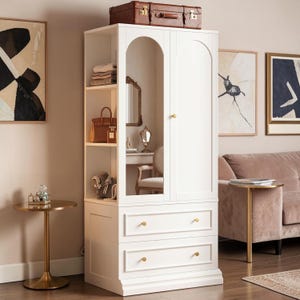 May include: A white wardrobe with an arched mirror and two drawers. The wardrobe has open shelving displaying a handbag, jewelry, and folded clothing. A brown suitcase sits on top. A small gold side table and a pink sofa are in the background.
