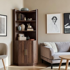 May include: A tall, dark wood corner cabinet featuring three open shelves filled with decorative vases, books, and small objects, above a lower storage section with two doors accented by vertical gold-toned handles and arched detailing.
