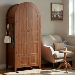 May include: A tall, wooden wardrobe with an arched top and two doors. The wardrobe has a drawer at the bottom and a section with hooks on the side. A white purse and a patterned tie hang from the hooks. A beige armchair and small wooden table are nearby.