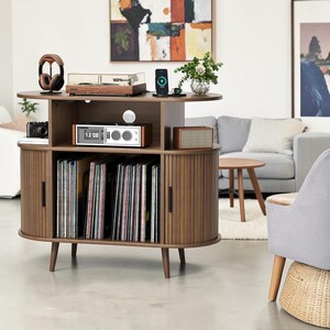 May include: A walnut-toned wooden media console featuring a top surface for a turntable, a middle shelf with a digital clock, and a lower section with vertical storage for vinyl records. The unit has two curved side cabinets with black handles.