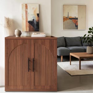 May include: A rectangular wooden cabinet with a medium brown finish, featuring two doors with vertical fluted paneling and arched detailing. The cabinet has two slim, vertical black metal handles centered on the doors for opening.
