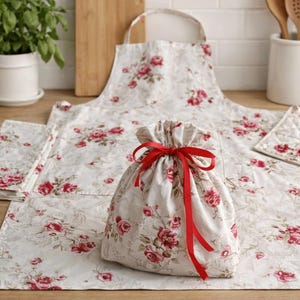 May include: A kitchen set featuring a floral pattern with red roses on a cream background. The set includes an apron, a drawstring bag tied with a red ribbon, a potholder, and a set of napkins. The items are arranged on a wooden surface.