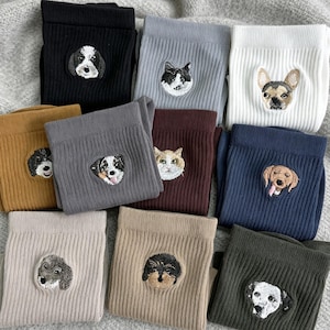 Könnte beinhalten: Eine Sammlung von zehn gerippten Stoffsocken in verschiedenen Farben wie Schwarz, Grau, Weiß, Senfgelb, Braun und Marineblau. Jede Socke verfügt über einen detaillierten gestickten Aufnäher mit einem Hunde- oder Katzengesicht.