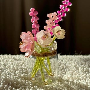 May include: A miniature glass jar filled with beaded flowers. The flowers are pink and white, with green stems. The jar is wrapped with twine. The background is a soft, blurred brown.