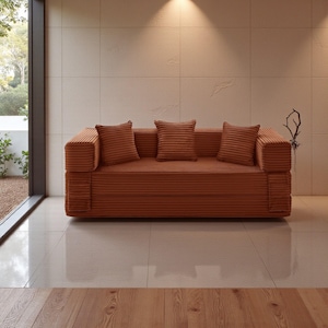 Könnte beinhalten: Ein rostfarbenes Cord-Sofa mit drei passenden Kissen. Das Sofa hat ein niedriges Profil und ein minimalistisches Design, ideal für einen modernen Wohnraum. Der Raum hat große Fenster und eine neutrale Farbpalette.