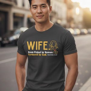 May include: Dark grey t-shirt featuring the word "WIFE" in gold, with a couple graphic. The text "Hand-Picked by Heaven. Favoured by God. PROVERBS 18:22" is below the word "WIFE".