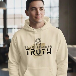 Puede incluir: Sudadera con capucha color crema con bolsillo delantero y capucha con cordón. La parte delantera presenta el texto "TRANSFORMED by TRUTH" en negro, con un gráfico de corazón y flecha arriba y "ROMANS 12:2" abajo. La sudadera es usada por una persona.