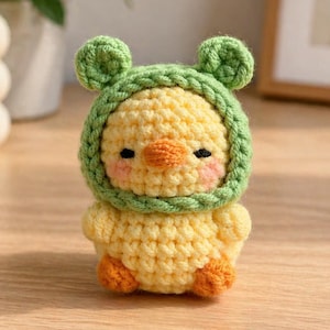 May include: A handmade crochet chick wearing a green hat with bear ears. The chick is yellow with an orange beak and feet, and pink cheeks. The background is a wooden surface with a blurred plant and a framed picture.