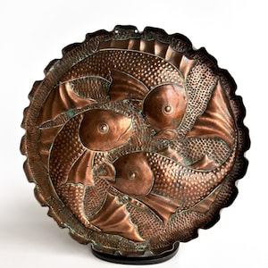 Puede incluir: Un plato redondo de cobre con borde festoneado que presenta un diseño en relieve de tres peces dispuestos en un patrón circular. La superficie tiene una textura martillada con áreas de oxidación verde entre los peces y los bordes.
