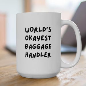 May include: White ceramic mug with a large handle. The mug has the text "WORLD'S OKAYEST BAGGAGE HANDLER" in black, bold letters. The mug is sitting on a wooden surface.