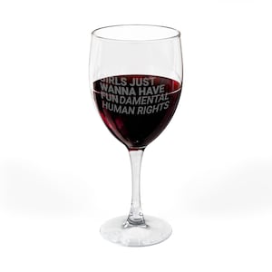 May include: A clear wine glass filled with red wine. The glass has the text "GIRLS JUST WANNA HAVE FUNDAMENTAL HUMAN RIGHTS" etched on the side. The glass sits on a white surface.