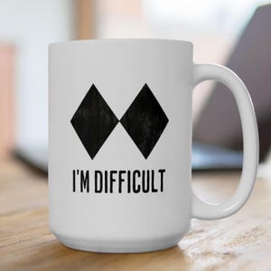 May include: White ceramic mug featuring two black diamond shapes and the words "I'M DIFFICULT" in black. The mug has a curved handle and is on a wooden surface.