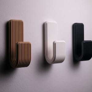 Könnte beinhalten: Drei Wandhaken in einer Reihe, jeder mit einer gebogenen J-Form. Die Haken sind braun, weiß und schwarz und haben eine gerippte Textur. Die Haken sind an einer hellgrauen Wand befestigt.
