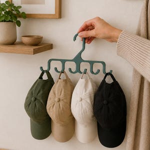 May include: A dark green plastic hanger with multiple hooks designed for organising accessories. Four baseball caps in olive green, tan, cream, and black are suspended from the hooks, showcasing a practical storage solution for headwear.