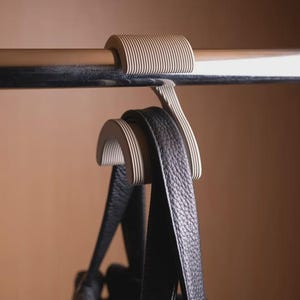 Könnte beinhalten: Ein beigefarbener, gerippter S-förmiger Kunststoffhaken, der über einer horizontalen Metallstange hängt. Der untere gebogene Teil des Hakens stützt den schwarzen Lederriemen einer Handtasche und hält diese sicher an der Stange.