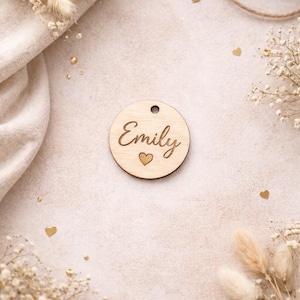 Könnte beinhalten: Ein runder Holzanhänger mit dem eingravierten Namen "Emily" in Schreibschrift und einem kleinen Herzsymbol. Der Anhänger hat ein Loch zum Aufhängen. Der Hintergrund ist beige mit dekorativen Elementen.