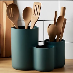 Könnte beinhalten: Ein tealfarbener Utensilienhalter mit drei Fächern, gefüllt mit Holzlöffeln und -spateln. Der Halter steht auf einer Holzoberfläche, im Hintergrund ein Schneidebrett und ein Glas mit Mehl. Die Küchenutensilien sind aus hellem Holz.