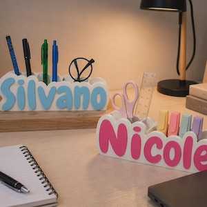 Peut inclure: Deux organisateurs de bureau blancs avec plusieurs fentes pour la papeterie. L'un porte le nom Silvano en lettres bleu clair et contient des stylos et des lunettes. L'autre porte le nom Nicole en lettres roses et contient des ciseaux, une règle et des surligneurs.