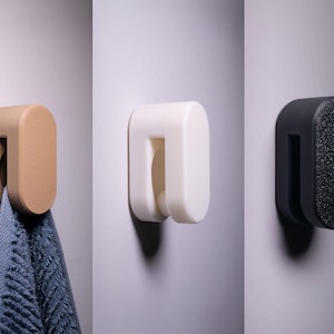 Könnte beinhalten: Drei Wandhaken in verschiedenen Farben: Beige, Weiß und Schwarz. Jeder Haken hat eine abgerundete rechteckige Form mit einer U-förmigen Öffnung. Ein blaues Handtuch hängt an dem beigefarbenen Haken. Der schwarze Haken hat eine strukturierte Oberfläche.