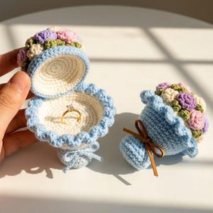 May include: Two crocheted ring boxes shaped like flower bouquets. One box is open, revealing a gold ring with a clear stone inside. Both feature light blue bases, ruffled edges, and clusters of small pink, purple, and cream-colored flowers.