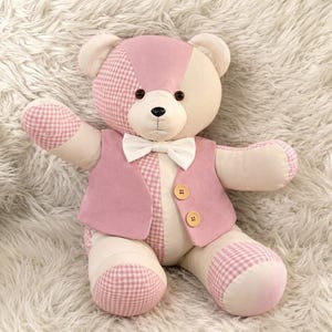 May include: A plush teddy bear featuring a patchwork design of cream and light pink fabric with a pink and white gingham pattern. The bear wears a solid pink vest with two light brown buttons and a white bow tie around its neck.