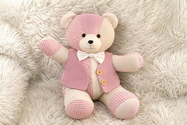 May include: A plush teddy bear featuring a patchwork design of cream and light pink fabric with a pink and white gingham pattern. The bear wears a solid pink vest with two light brown buttons and a white bow tie around its neck.