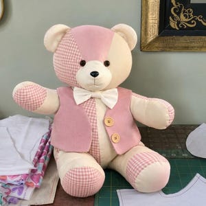 May include: A plush teddy bear constructed from patchwork fabric in shades of cream and light pink, featuring a gingham pattern on the ears, arms, and legs. The bear wears a solid pink vest with two wooden buttons and a white bow tie at the neck.