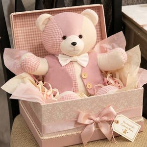 May include: A plush teddy bear with pink and cream patchwork fur, wearing a pink vest and a white bow tie, seated inside a decorative pink gift box lined with tissue paper. The box features a pink ribbon bow and a small tag with text.