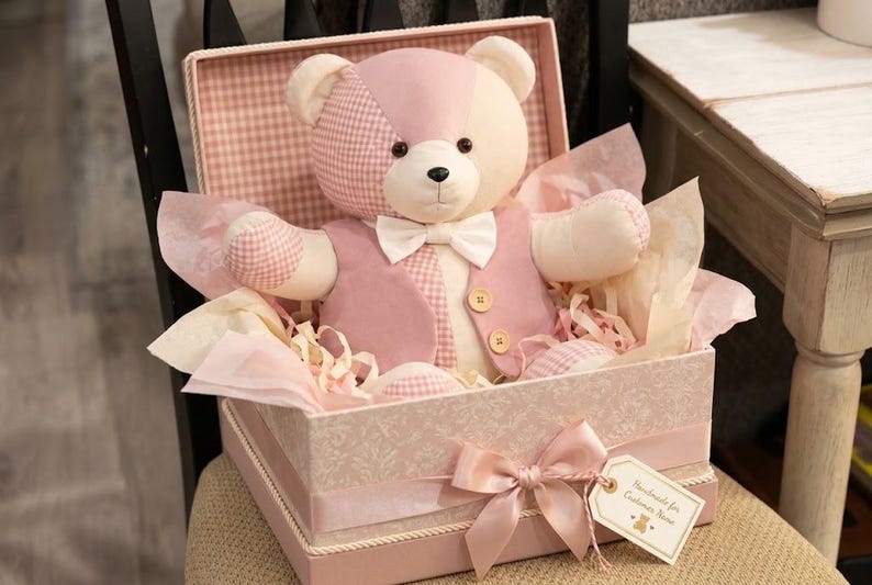 May include: A plush teddy bear with pink and cream patchwork fur, wearing a pink vest and a white bow tie, seated inside a decorative pink gift box lined with tissue paper. The box features a pink ribbon bow and a small tag with text.