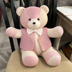 May include: A plush teddy bear featuring a patchwork design of cream and pink fabric, including sections with a pink and white gingham pattern. The bear wears a pink vest with two light brown buttons and a white bow tie at the neck.