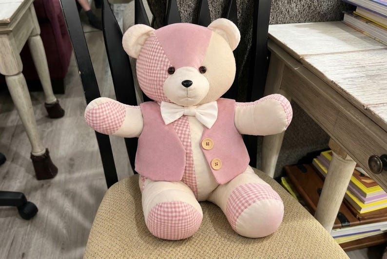 May include: A plush teddy bear featuring a patchwork design of cream and pink fabric, including sections with a pink and white gingham pattern. The bear wears a pink vest with two light brown buttons and a white bow tie at the neck.