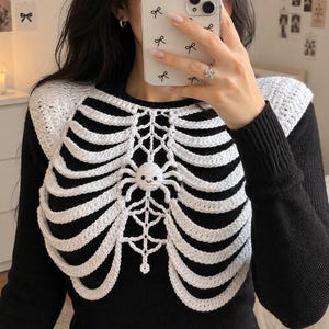 May include: A white crocheted rib cage overlay worn over a black long-sleeved shirt. The white yarn piece features a detailed skeletal rib structure with a small, central crocheted spider resting on a web design positioned at the sternum area.
