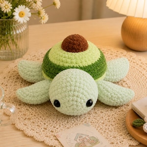 May include: A crocheted avocado turtle toy. The turtle has a light green body, four flippers, and a brown pit. The shell is green and striped. The toy is on a beige crocheted doily.