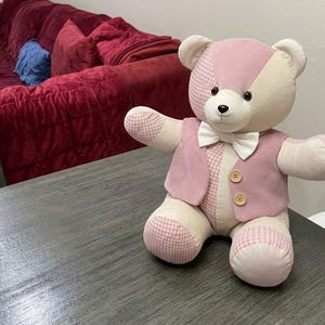 May include: A stuffed teddy bear constructed from patchwork fabric in shades of cream and light pink, featuring a mix of solid and gingham patterns. The bear wears a matching light pink vest with two wooden buttons and a white bow tie at the neck.