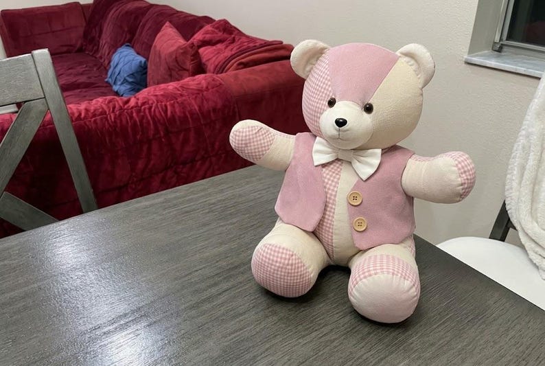 May include: A stuffed teddy bear constructed from patchwork fabric in shades of cream and light pink, featuring a mix of solid and gingham patterns. The bear wears a matching light pink vest with two wooden buttons and a white bow tie at the neck.