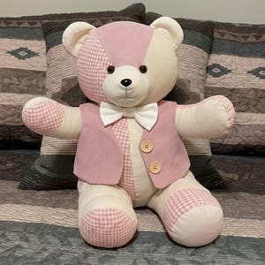 May include: A plush teddy bear featuring a patchwork design of cream and pink gingham fabric. The bear wears a pink vest with two light brown wooden buttons and a white bow tie. It has black plastic eyes and a black stitched nose and mouth.