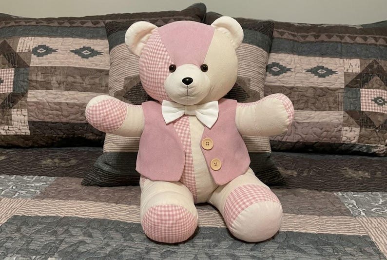 May include: A plush teddy bear featuring a patchwork design of cream and pink gingham fabric. The bear wears a pink vest with two light brown wooden buttons and a white bow tie. It has black plastic eyes and a black stitched nose and mouth.