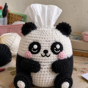 May include: A crochet tissue box cover shaped like a panda with a white face, black ears, black limbs, and pink cheeks. The panda has embroidered black eyes with white highlights and a small black nose and mouth. A white tissue protrudes from the top.