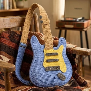 May include: A crochet guitar-shaped bag in light blue with a yellow pickguard and dark grey accents. The bag features a long, adjustable tan crochet strap with two dark buttons near the top and a metal buckle for length adjustment.