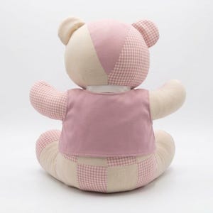 May include: A stuffed teddy bear featuring a patchwork design of cream-colored fabric and pink gingham fabric. The bear is wearing a solid pink vest over its torso and has a white ribbon collar around its neck, viewed from the back.